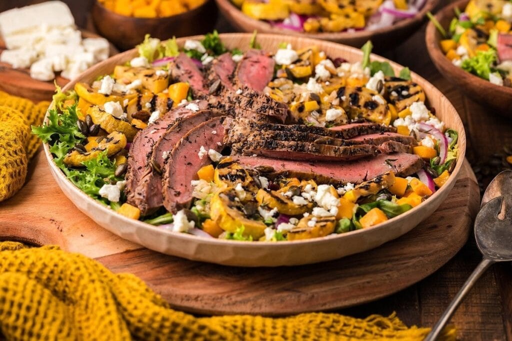 A wooden bowl filled with a mixed salad topped with sliced grilled steak, roasted squash, crumbled feta cheese, and diced orange vegetables sits on a wooden board next to a yellow cloth—an inspiration for delicious steak recipes.