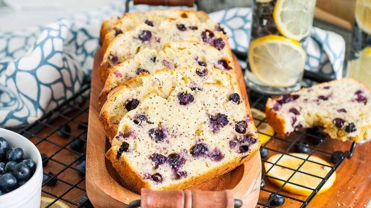 Slices of lemon blueberry poppy seed loaf on a cutting board and a bowl of fresh blueberries with glasses filled with blueberries and lemon slices in the background. 