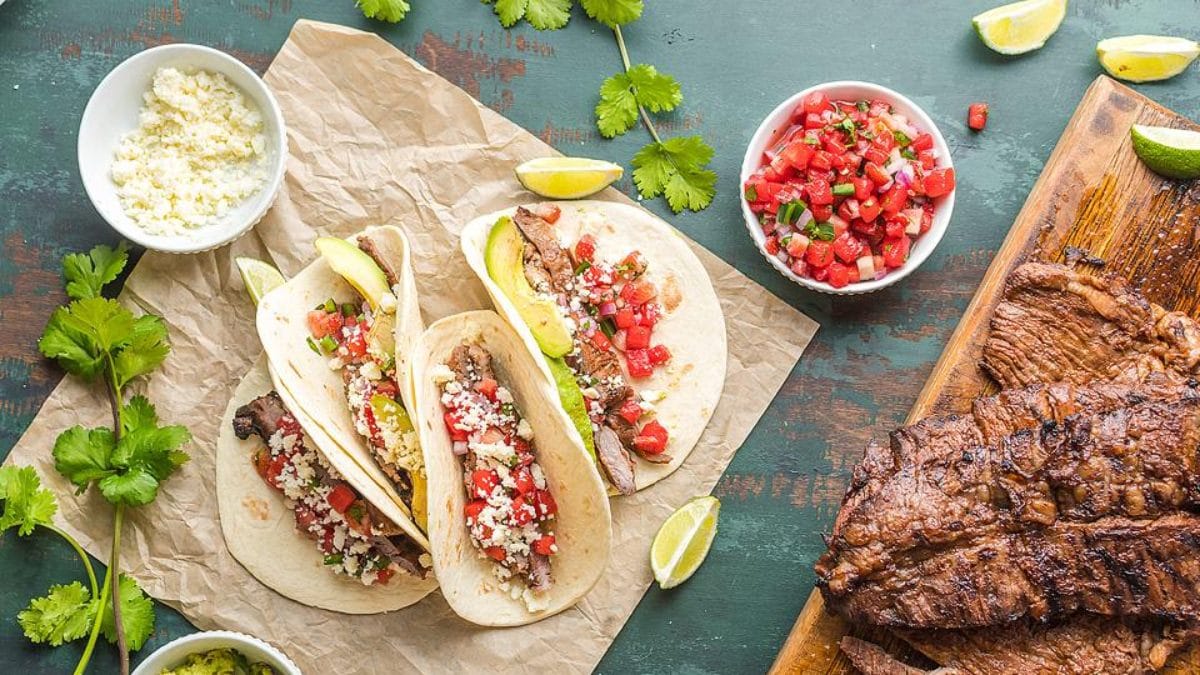 Three marinated grilled flank steak tacos with tomatoes, cheese, and avocado slices.