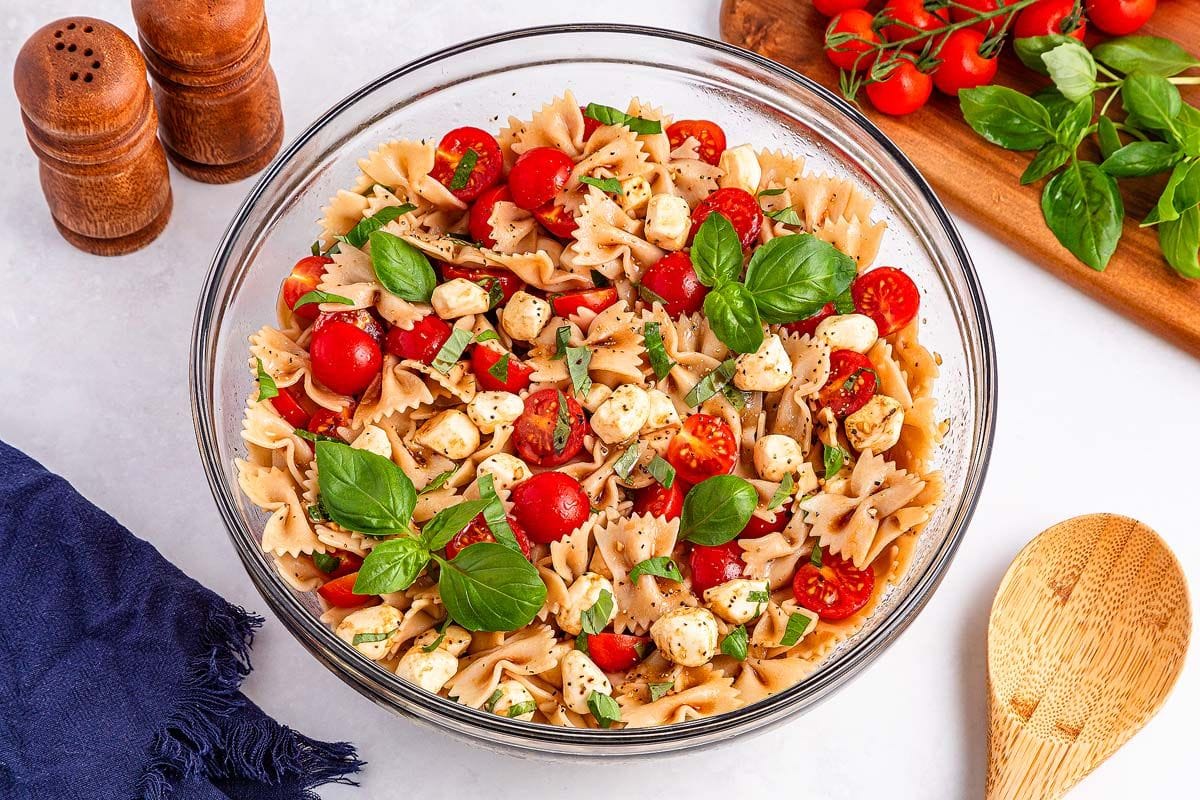 Caprese pasta salad in a white dish on a wooden round board, and a dish of dressing and bread in the background.