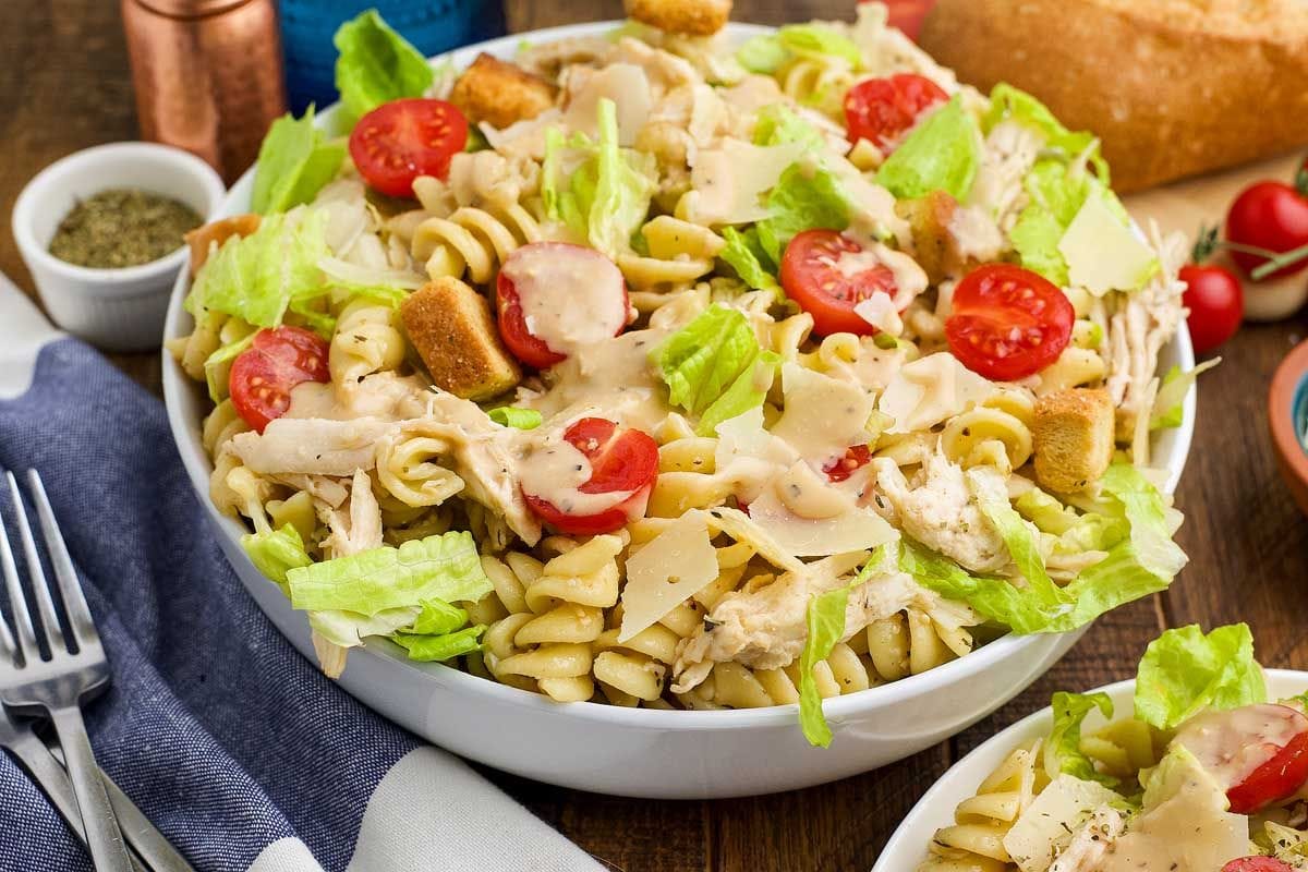 Chicken Caesar pasta salad in a white bowl with a wooden spoon lifting a bite.