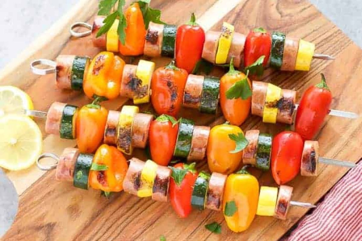 Four chicken sausage skewers with vegetables on a wooden cutting board.