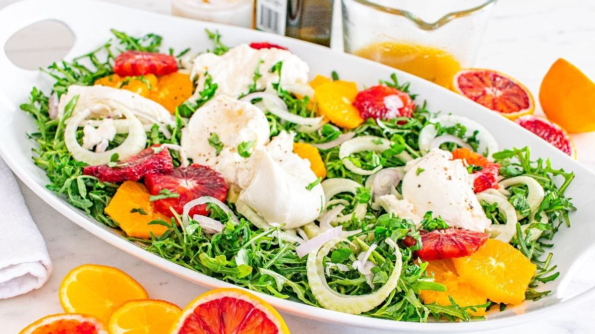 Fennel orange salad on a blue plate on a table with two forks, orange slices, a small bowl of dressing, and more salad in a serving dish.