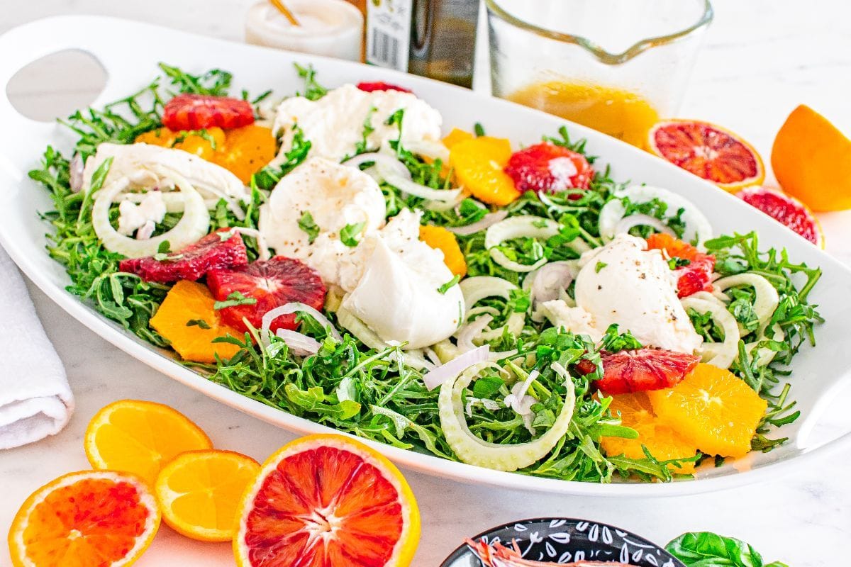 Fennel orange salad on a blue plate on a table with two forks, orange slices, a small bowl of dressing, and more salad in a serving dish.